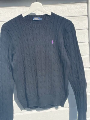 Mörkblå kabelstickad tröja Polo Ralph Lauren - Kabelstickad mörkblå tröja från Polo Ralph Lauren i 100% bomull. Klassisk rund halsringning och lång ärm. Tröjan har den ikoniska rosa broderade loggan på bröstet och ribbade muddar vid ärmslut och nederkant. Perfekt för en stilren och avslappnad look.