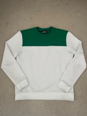 Vit och grön sweatshirt J.Lindeberg - Snygg sweatshirt från J.Lindeberg i vitt med grön panel upptill och diskret logga på bröstet. Tröjan har rund halsringning och långa ärmar. Perfekt för en avslappnad och sportig stil.