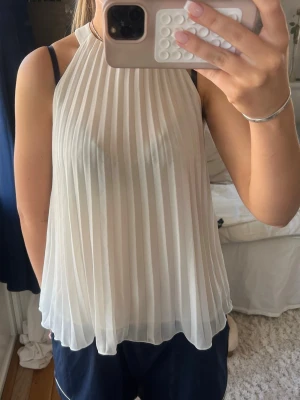 Vit plisserad ärmlös topp - Snygg vit ärmlös topp med plisserat tyg och hög hals. Luftig och lätt modell som ger en elegant känsla. Perfekt att matcha med jeans eller kjol för en stilren look. Står xs men passar S/M också.
