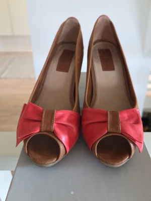 Bruna pumps med röd rosett - Snygga pumps i brunt skinn med öppen tå och klack. Framtill sitter en stor röd rosett som ger skorna en cool retrovibe. Klacken är träfärgad och skon har en mjuk innersula för extra komfort. Perfekta för dig som vill sticka ut lite extra.
