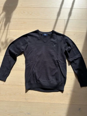 Gant svart tröja - Klassisk svart stickad tröja från Gant med V-ringning och diskret logga broderad på bröstet. Tillverkad i mjuk bomull som känns skön mot huden. Perfekt att bära över en skjorta eller t-shirt för en clean look.