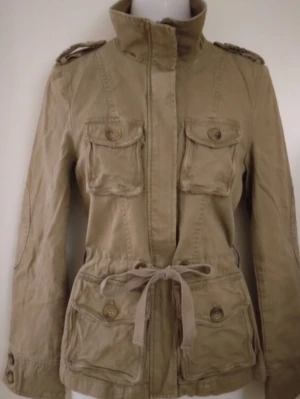 Beige parkas / sommarjacka / vårjacka / cargojacka / utilityjacka. Strlk: XS. Felfri. - Beige parkas / sommarjacka / vårjacka / cargojacka / utilityjacka. Strlk: XS.  Fint skick, knappt använd, felfri. Kraftigare bomullstyg samt fint äppelmönstrat foder inuti. Flera fickor. Stängs med dragkedja och har knytband i midjan. FRAKT TILLKOMMER.