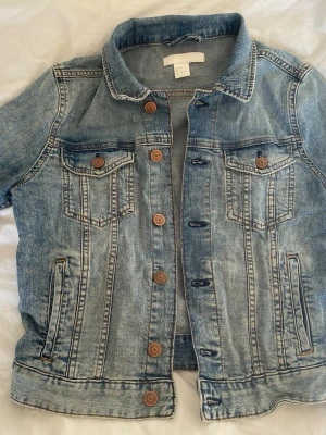 Blå klassisk jeansjacka - Säljer en blå jeansjacka från H&M med klassisk design, bröstfickor med knappar. Jackan har bruna knappar framtill. Inga defekter