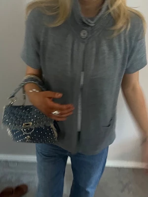 Grå stickad kofta med kort ärm - Grå stickad kofta med korta ärmar och hög krage med stor knappdetalj. Koftan har två fickor framtill och ribbade kanter. Perfekt att slänga över en t-shirt för en chill look.