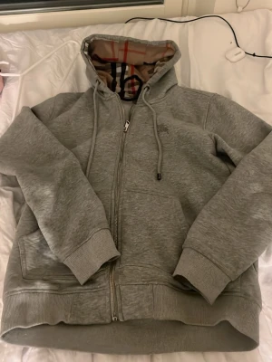 Grå zip hoodie från Burberry - Säljer en grå hoodie från Burberry med dragkedja och klassiskt rutigt foder i huvan. Hoodien har ribbade muddar, fickor framtill och justerbar dragsko i huvan. Perfekt för dig som gillar stilrena och exklusiva plagg med en twist. Storlek M men passar S
