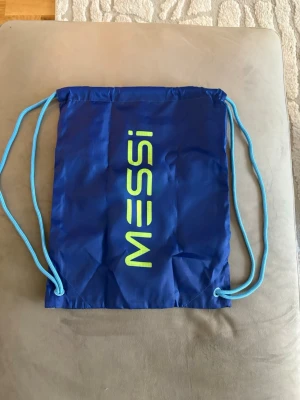Blå Messi gympåse med snören - Snygg blå gympåse med ljusblå snören och tryck i neongult med texten 'Messi' på ena sidan och Messi-logga på andra. Tillverkad i lätt och slitstarkt syntetmaterial, perfekt för träning eller skola.