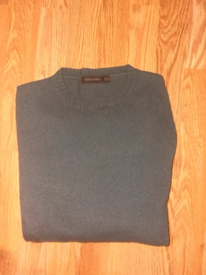 Mörkgrön Kaschmir Tröja Oscar Jacobson - En stilren mörkgrå kaschmir tröja från Oscar Jacobson med rund halsringning och långa ärmar. Tröjan har ribbstickade muddar vid ärmslut och nederkant. Perfekt för lager-på-lager och enkel att matcha med olika outfits. Det är en kaschmir- och ullmix.