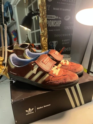Adidas x Wales Bonner röda sneakers - Unika sneakers från Adidas i samarbete med Wales Bonner. Modellen har en röd ovandel i mocka med beigea detaljer och snörning, samt bred kardborre över vristen. Klassiska Adidas-ränder på sidan och svart sula. Perfekt för dig som vill sticka ut.