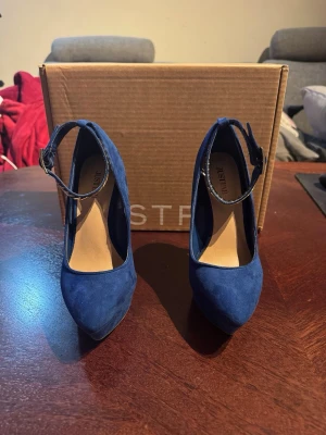 Blåa pumps med ankelrem från JustFab - Snygga blåa pumps från JustFab med spetsig tå och hög smal klack. Skorna har en elegant ankelrem med spänne och är tillverkade i ett mockaliknande material. Perfekta för dig som vill sticka ut med färg och stil.