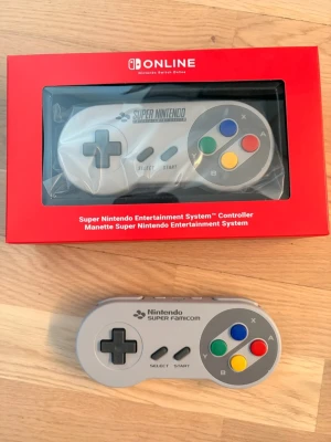 Nintendo Super Famicom handkontroll - Klassisk Super Famicom handkontroll från Nintendo med grått plastskal och färgglada knappar i blått, grönt, gult och rött. Ergonomisk retrodesign med styrkors, Select- och Start-knappar. Perfekt för Switch Online och nostalgiska spelkvällar.