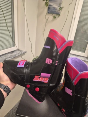 Salomon Pjäxor HTC 520 svarta skidkängor 44 28.5cm - Säljer ett par retro Salomon HTC 520 skidkängor i svart plast med detaljer i lila, rosa och rött. Kängorna har en futuristisk design med spännen och vadderad insida i lila. Perfekta för dig som vill sticka ut i backen i vinter!