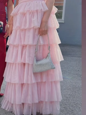 Ljusrosa volangklänning maxilängd - Säljer en ljusrosa maxiklänning med flera lager volanger och djup rygg. Klänningen har ärmlös design och dragkedja bak. Tillverkad i ett tunt, flowy tyg som ger en romantisk och drömmig look. Perfekt för bal eller student.