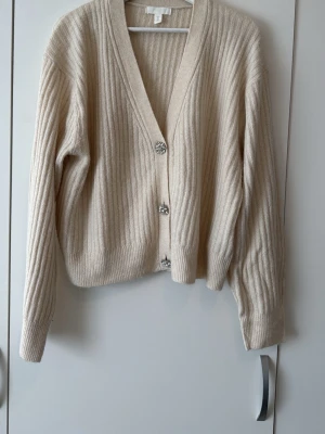 Beige ribbad kofta med pärlknappar - Snygg beige ribbstickad kofta med v-ringning och dekorativa pärlknappar. Koftan har långa ärmar och en avslappnad passform, perfekt att slänga över en t-shirt eller topp. Materialet känns mjukt och skönt mot huden.