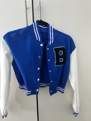 Blå och vit bomberjacka med B - Snygg blå bomberjacka med vita ärmar och stora bokstaven B på bröstet. Jackan har ribbade muddar och krage med vita ränder, tryckknappar framtill och klassisk college-look. Perfekt för dig som vill sticka ut med en sportig och trendig stil.