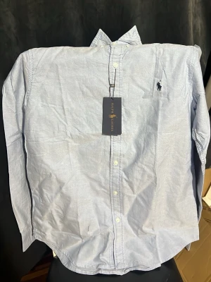 Ljusblå skjorta från Ralph Lauren - Klassisk ljusblå skjorta från Ralph Lauren med button-down krage och broderad logga på bröstet. Skjortan har långa ärmar och knäppning framtill. Perfekt till jeans eller chinos för en clean och stilren look.