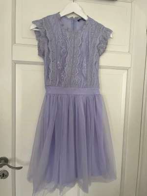 Jättesnygg spetsklänning - Söt ljuslila klänning med spets upptill och volangig kjol i tunn tyll. Klänningen har korta ärmar och rund halsringning. Perfekt för dig som gillar romantisk stil och mjuka pasteller.