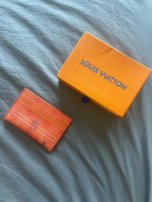 Orange kortfodral - Säljer ett orange kortfodral med grafiskt tryck och flera kortfack. Tillverkad i tåligt läder med exklusiv känsla och klassisk design. Perfekt för dig som vill ha en snygg och praktisk accessoar från ett ikoniskt märke.
