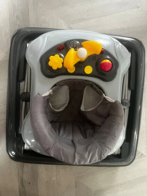 Grå gåstol med leksakspanel - Säljer en grå gåstol med mjuk sits och en leksakspanel framtill. Panelen har färgglada knappar och detaljer i gult, rött och vitt som gör det roligt för barnet att utforska. Perfekt för små barn som vill träna på att gå och leka samtidigt.