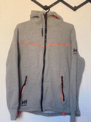Grå Helly Hansen hoodie med dragkedja - Snygg grå hoodie från Helly Hansen med hel dragkedja och orange detaljer på fickor och bröst. Märket syns på både ärm och huva. Hoodien har två fickor med dragkedja och justerbar huva. Perfekt för chill dagar eller när du vill ha en sportig look.