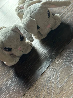 Tofflor med kaninmotiv och öron - Supersöta tofflor i beige plysch med kaninansikte, broderade ögon, rosa nos och långa mjuka öron. Insidan är ljusrosa och baktill finns en fluffig svansboll. Perfekta för att hålla fötterna varma hemma och ger en gullig look.