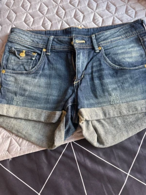 Blå jeansshorts från C Denim - Snygga blå jeansshorts från C Denim med uppvikta benslut och klassiska fem fickor. Shortsen har guldiga knappar och detaljer samt slitningar framtill för en cool look. Perfekta för varma dagar och enkel att matcha med olika toppar.