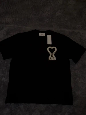 Svart t-shirt från Ami Paris - Svart t-shirt från Ami Paris med ikonisk hjärtlogga i vitt på bröstet. Klassisk passform och rund halsringning. Perfekt till jeans eller shorts för en clean och stilren look. Skirv vid intresse eller om du har några funderingar 