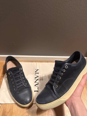 Mörkblå sneakers från Lanvin - Snygga mörkblå sneakers från Lanvin med ovandel i mocka och läder, vita gummisulor och klassisk låg profil. Skorna har metallfärgade öljetter och matchande mörkblå skosnören. Perfekta för dig som gillar stilrena och exklusiva sneakers.