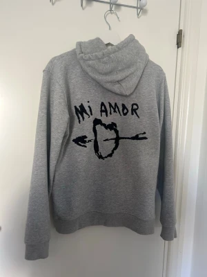 Grå hoodie med tryck 'Mi Amor' - Säljer en grå hoodie med huva och magficka. På ryggen finns ett svart tryck med texten 'Mi Amor' och ett hjärta genomborrat av en pil. Hoodien har dragsko i huvan och är gjord i mjukt sweatshirtmaterial, perfekt för en chill och avslappnad stil.