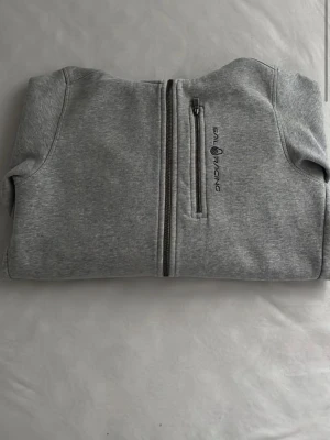 Grå hoodie från Sail Racing - Snygg grå hoodie från Sail Racing med dragkedja framtill och en extra bröstficka med zip. Tillverkad i mjukt sweatshirtmaterial och har en broderad logga på bröstet samt på huvan. Perfekt för en avslappnad och sportig stil.