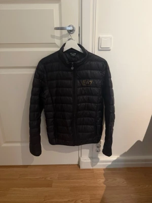 Svart dunjacka från EA7 Emporio Armani - Snygg svart dunjacka från EA7 Emporio Armani med quiltad design och guldfärgad logga på bröstet. Jackan har dragkedja framtill och står för en clean och modern look. Perfekt för kyliga dagar när du vill hålla stilen.
