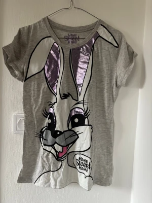 Grå pyjamas med kanintryck - Söt pyjamas med grå t-shirt med stort kanintryck och metallic-lila detaljer i öronen samt texten 'Home Sweet Home'. Tillhörande byxor i grått med lila och vita rutmönster samt lila midjeband. Perfekt för mysiga hemmakvällar.