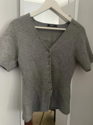 Grå kortärmad kofta från Gina Tricot - Stilren grå kofta från Gina Tricot med korta ärmar och v-ringning. Koftan har knappar framtill och är gjord i ett mjukt, stickat material som sitter snyggt på kroppen. Perfekt att matcha med jeans eller kjol för en enkel och trendig look.