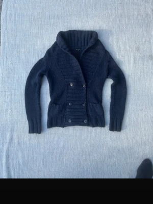 Mörkblå dubbelknäppt kofta - Snygg mörkblå stickad kofta med dubbelknäppning och bred krage. Koftan har långa ärmar och ribbade muddar vid ärmslut och nederkant. Perfekt för att hålla sig varm och samtidigt se stilren ut.
