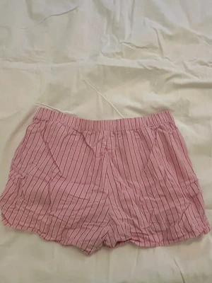 Rosa pyjamasshorts med ränder - Säljer ett par rosa pyjamasshorts med tunna vertikala vita ränder. Shortsen har elastisk midja och är gjorda i ett lätt och mjukt tyg, perfekta för att chilla hemma eller sova i. De har en avslappnad passform och känns riktigt bekväma.