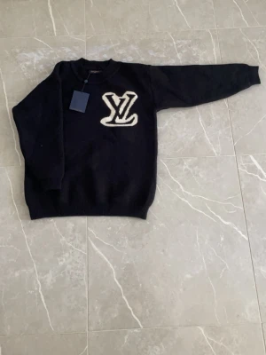 Svart stickad tröja med LV-logga - Säljer en svart stickad tröja från Louis Vuitton med stor vit LV-logga framtill och baktill. Tröjan har rund halsringning, långa ärmar och ribbade muddar vid ärmslut och nederkant. Perfekt för dig som gillar exklusiva märken och stilren design.