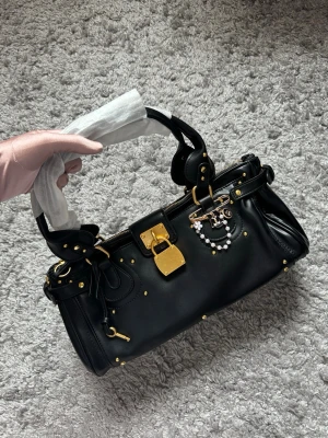 Svart handväska med gulddetaljer - Snygg svart handväska med guldfärgade metalldetaljer och dekorativa nitar. Väskan har ett robust handtag, hänglås framtill och en charm med pärlor och logga. Perfekt accessoar för dig som gillar lyxig och edgy stil.