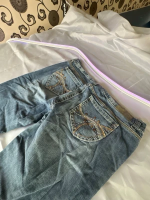 Blå jeans med broderade fickor Y2k premier  - Snygga blå jeans med raka ben och detaljerade broderier på bakfickorna. Jeansen har klassisk femficksmodell och är gjorda i denim med en lätt tvättad look. Perfekta för dig som vill ha något extra på fickorna och gillar en avslappnad stil.