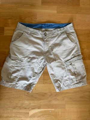 Beige cargoshorts med fickor - Ett par beige cargoshorts från Etirel med flera praktiska fickor på sidorna och bak. Klassisk passform med bälteshällor och knappstängning. Perfekta för sommardagar.