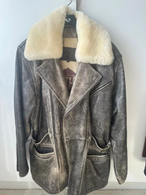 Grå/brun pälsjacka med stor krage - Snygg grå/brun pälsjacka med stor vit furpäls-krage. Jackan har en vintage-look, dragkedja, stora fickor och markerad midja. Perfekt för dig som vill sticka ut med en cool och unik stil.