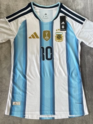 Argentina matchtröja Adidas nr 10 - Säljer en officiell Argentina fotbollströja från Adidas med klassiska blå och vita ränder, nummer 10 på bröstet och FIFA World Champions 2022 märke. Tröjan har AFA logga, guldiga Adidas detaljer. Helt oanvänd med prislapp! 
