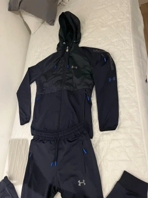 Svart träningsset från Under Armour - Snyggt svart träningsset från Under Armour med matchande hoodie och byxor. Setet har blå detaljer på dragkedjor och snören, samt logga på bröstet och benet. Jackan har huva och dragkedja framtill, byxorna har resår i midjan och praktiska fickor.