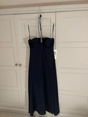 Mörkblå långklänning med knyte runt nacken - Elegant mörkblå långklänning med band man knyter runt nacken. Klänningen har en figurnära överdel och faller snyggt ner mot golvet. Perfekt för speciella tillfällen när du vill känna dig extra fin. Helt ny med alla lappar kvar! Pris kan diskuteras vid snabb affär.