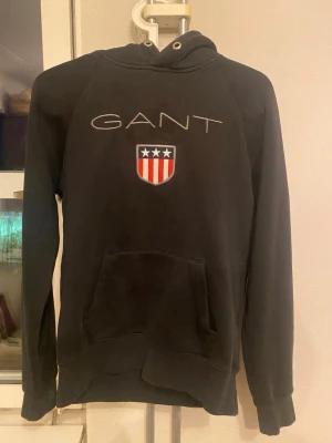 Svart hoodie från GANT med logga - Svart hoodie från GANT med broderad logga och sköld på bröstet. Klassisk känguruficka framtill och huva med dragsko. Mjuk bomullskänsla och avslappnad passform, perfekt för en chill stil.