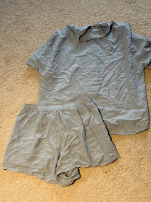 Ljusblåa pyjamas-set i linne - Mysigt ljusgrått pyjamas-set med kortärmad skjorta och matchande shorts. Skjortan har knappar framtill och en bröstficka, medan shortsen har resår i midjan för extra komfort. Tillverkad i ett tunt, luftigt linnematerial som känns svalt mot huden.