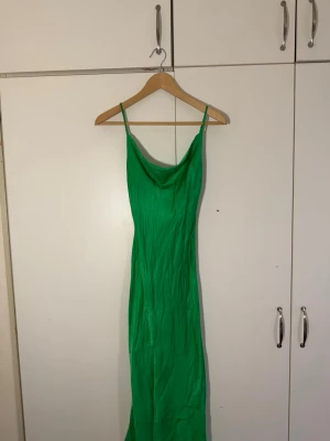 Grön långklänning med smala axelband i silkesmaterial  - Snygg grön långklänning med smala axelband, draperad urringning och en liten slits nertill. Klänningen har en slank passform och är tillverkad i ett glansigt material som ger en lyxig känsla. Perfekt för dig som vill sticka ut med en färgstark look.