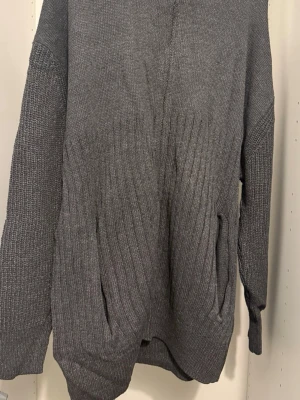 Grå stickad tröja med fickor - Mysig grå stickad tröja med långa ärmar och två praktiska fickor framtill. Tröjan har en enkel design med diskret ribbstickat mönster nedtill och vid ärmsluten. Perfekt för lager-på-lager och en avslappnad stil.