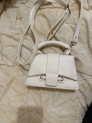 Vit minihandväska med axelrem - Snygg vit minihandväska i strukturerat konstläder med både handtag och justerbar axelrem. Väskan har ett dekorativt spänne framtill och guldfärgade detaljer. Perfekt för dig som gillar trendiga accessoarer.
