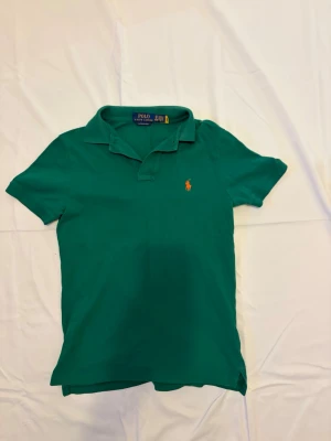 Grön pikétröja från Polo Ralph Lauren - Grön piké från Polo Ralph Lauren i modellen Custom Slim Fit. Tröjan är i gott skick utan synliga defekter, fläckar eller slitage. Detaljer: 	•	Märke: Polo Ralph Lauren 	•	Modell: Custom Slim Fit Piqué Polo 	•	Färg: Grön med orange inbrodererad pony-logga 	•	Storlek: XS men passar S 	•	Material: Bomull piqué 	•	Passform: Slim/tajt Skick: Gott — inga defekter.                                          Modellen: 178 cm
