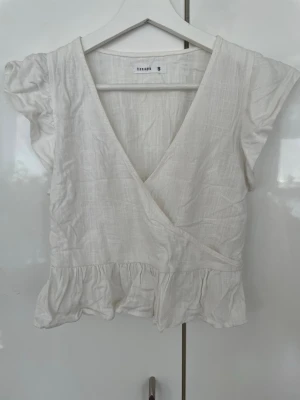 Vit linnetopp från terranova - Söt vit linnetopp från terranova med volangkant nedtill och korta, lätt puffade ärmar. Toppen har v-ringning och smock i ryggen för en bekväm passform. Perfekt till sommaren och enkel att styla med jeans eller kjol.