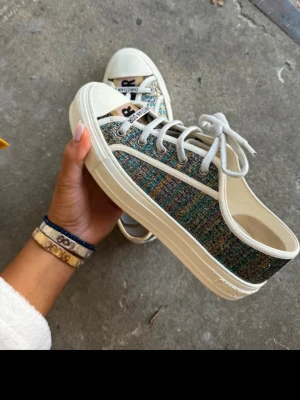 Glittriga låga sneakers från Dior - Säljer ett par unika sneakers från Christian Dior med glittrig, flerfärgad textil och vita detaljer. Skorna har vita snören, klassisk tåhätta och låg profil. Perfekta för dig som vill sticka ut med exklusiva och trendiga skor. Går bra med att komma med lägre prisförslag 